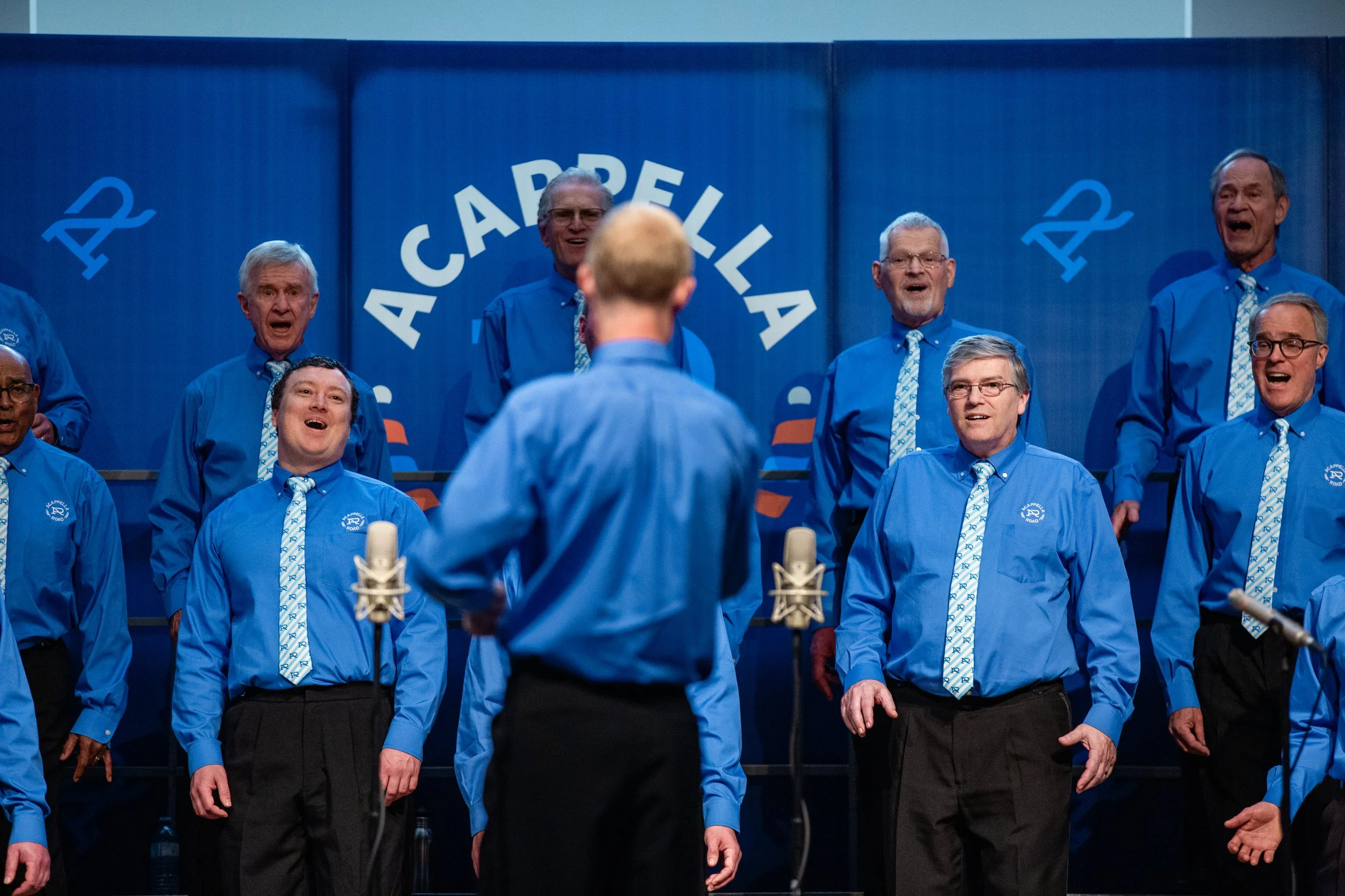 Members of Acappella Road performing onstage in blue shirts and patterned ties. Director Adrian Smith is positioned in the foreground in front of them, shot from behind while directing a song.