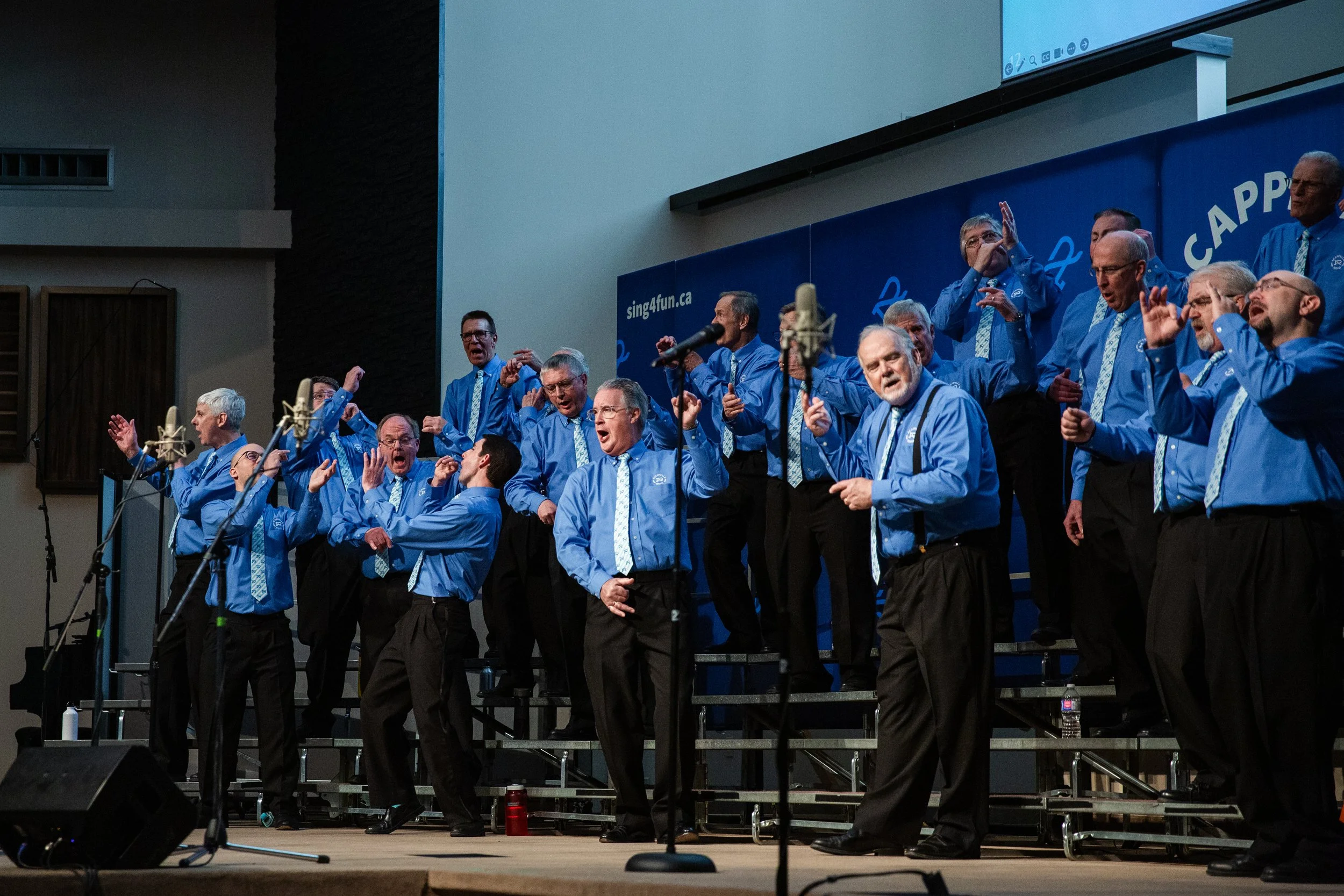 Acappella Road members performing in concert, dressed in blue uniforms with patterned ties. They are facing in all directions, pantomiming playing various instruments in a jazz band.