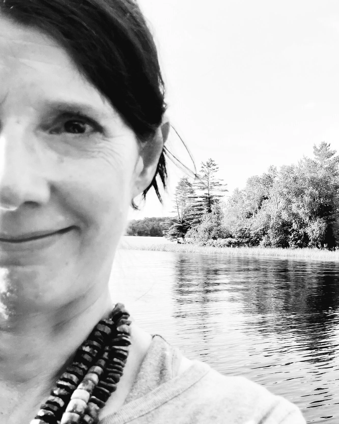 A woman with dark hair and a beaded necklace, smiling near a body of water with trees in the background, in black and white.