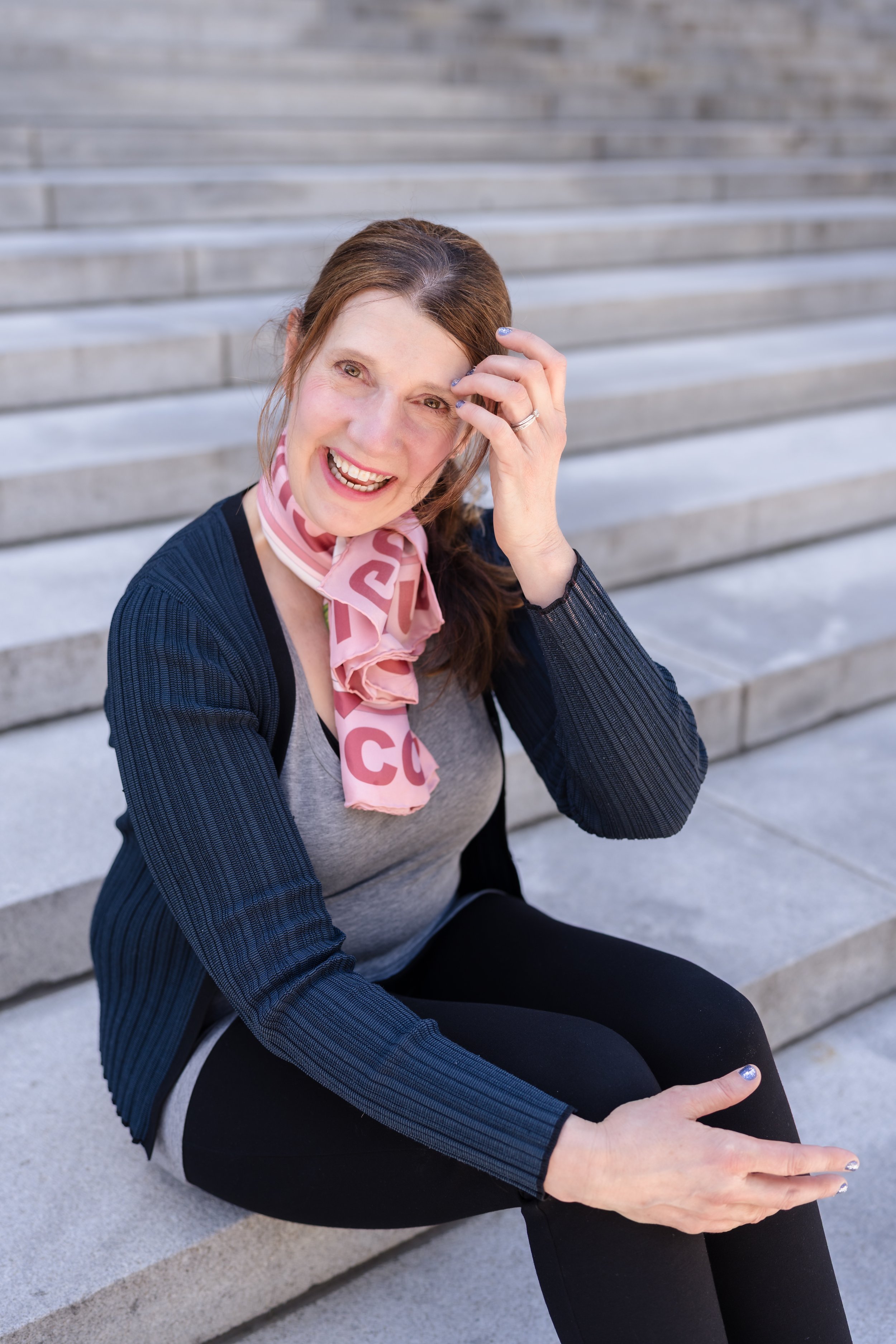 A woman with brown hair sitting on outdoor steps, smiling, touching her forehead, wearing a pink scarf, gray shirt, black pants, and a blue jacket.