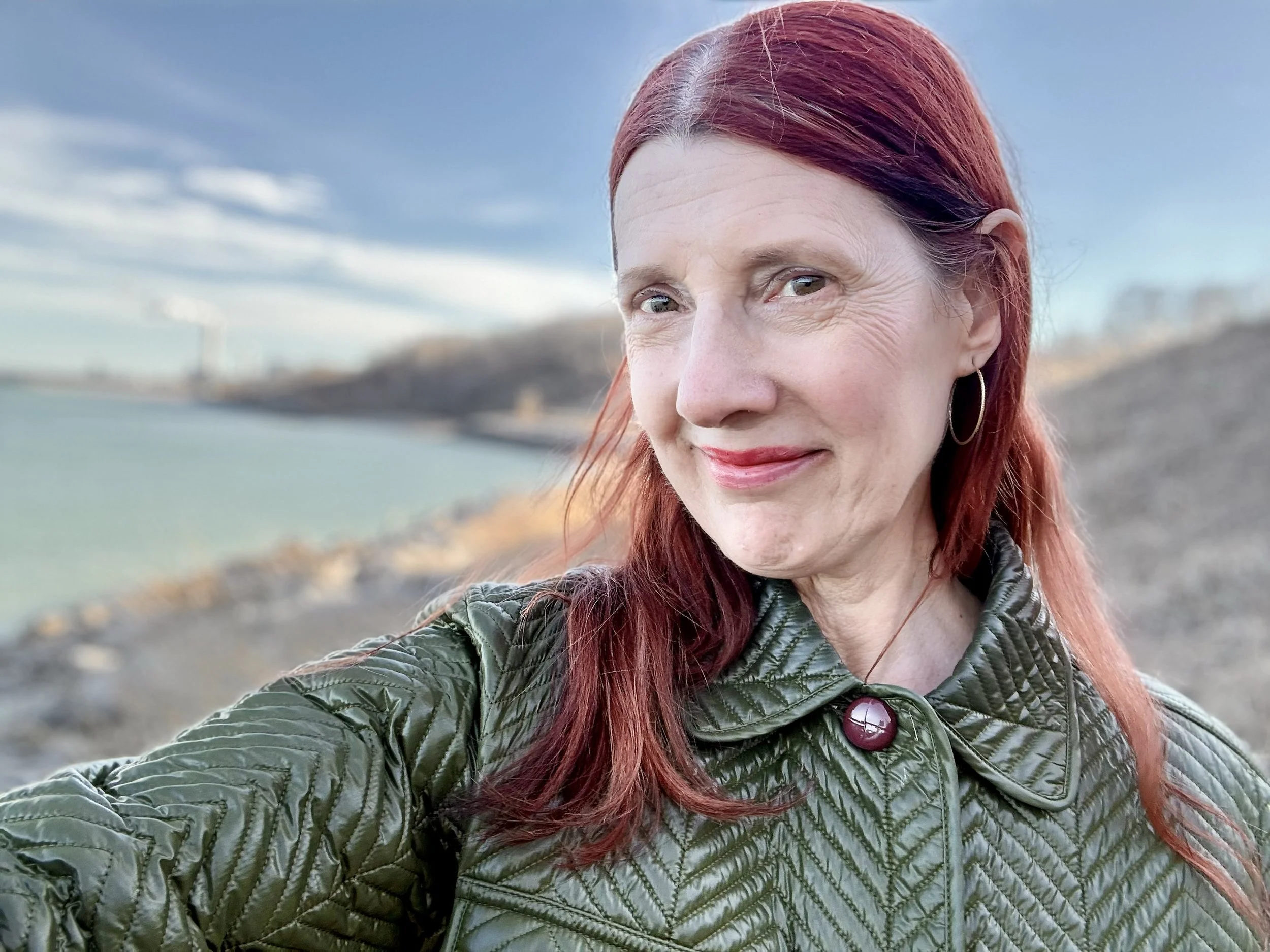 A woman with red hair smiling outdoors, wearing a green quilted jacket with a button, with a blurred background of water and sky.