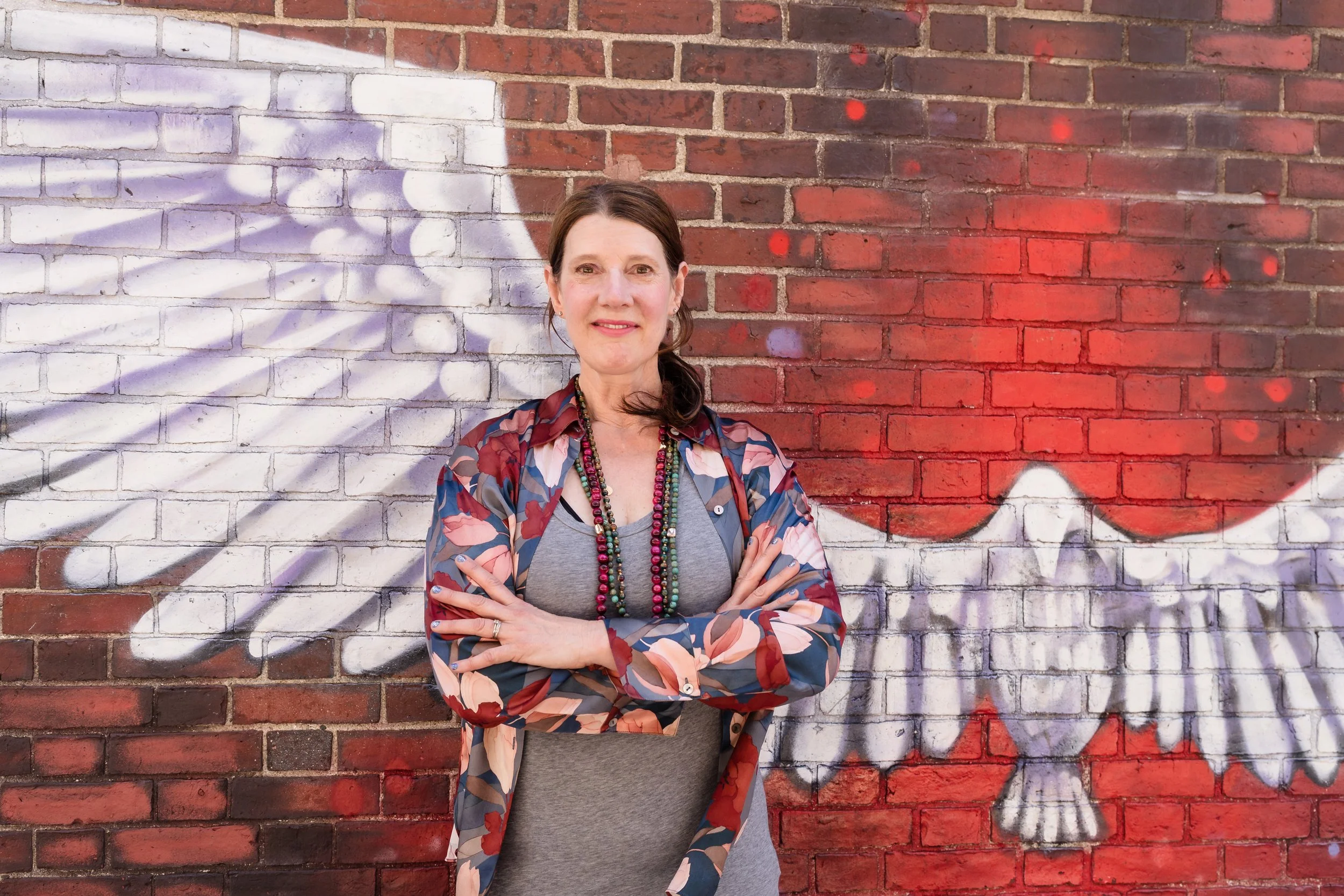 A woman with brown hair pulled back, wearing a gray shirt, layered necklaces, and a colorful floral jacket, stands with arms crossed in front of a brick wall that features white and purple graffiti.