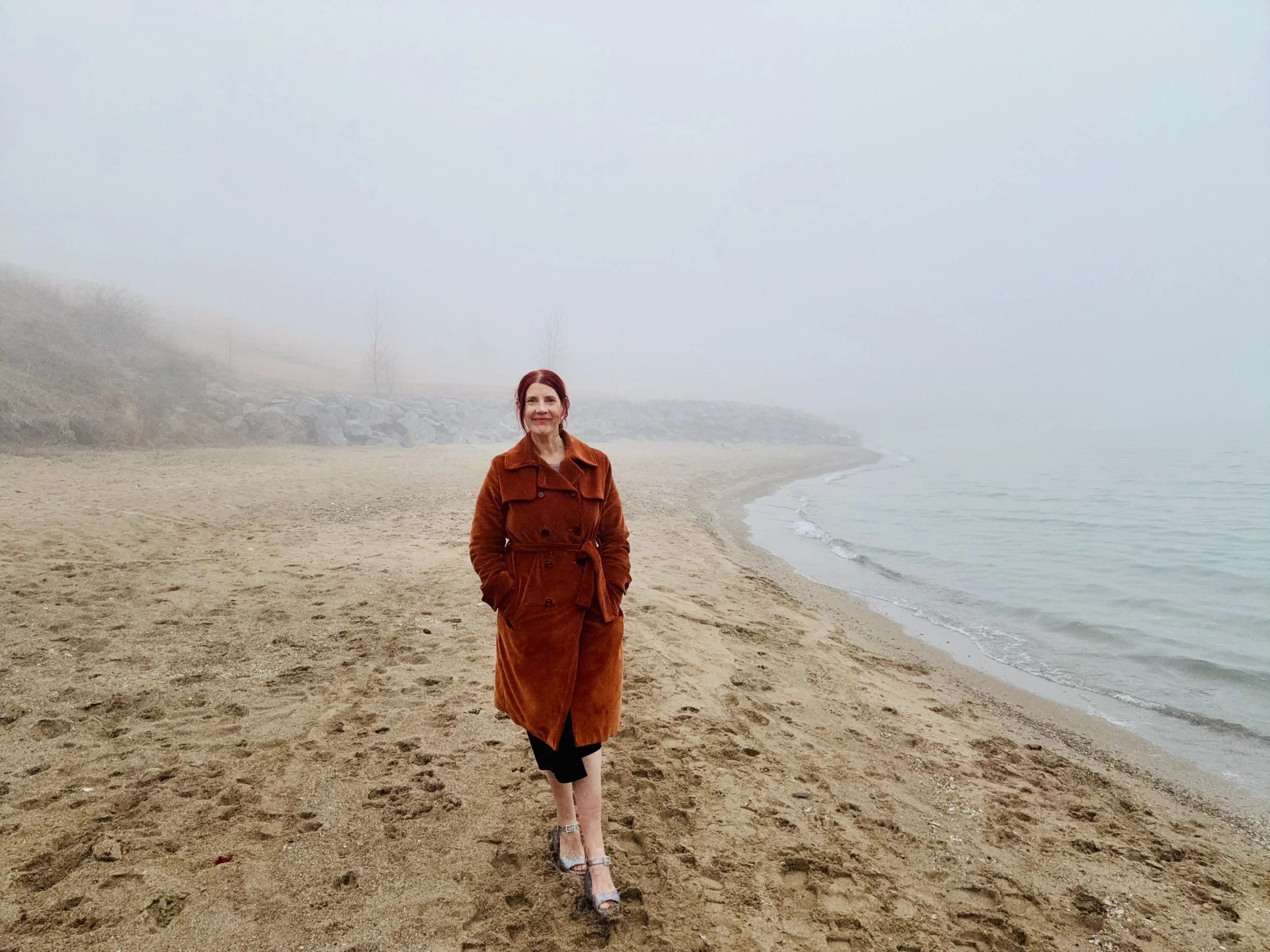 Woman with red hair wearing an orange coat and black skirt walking on a foggy sandy beach.