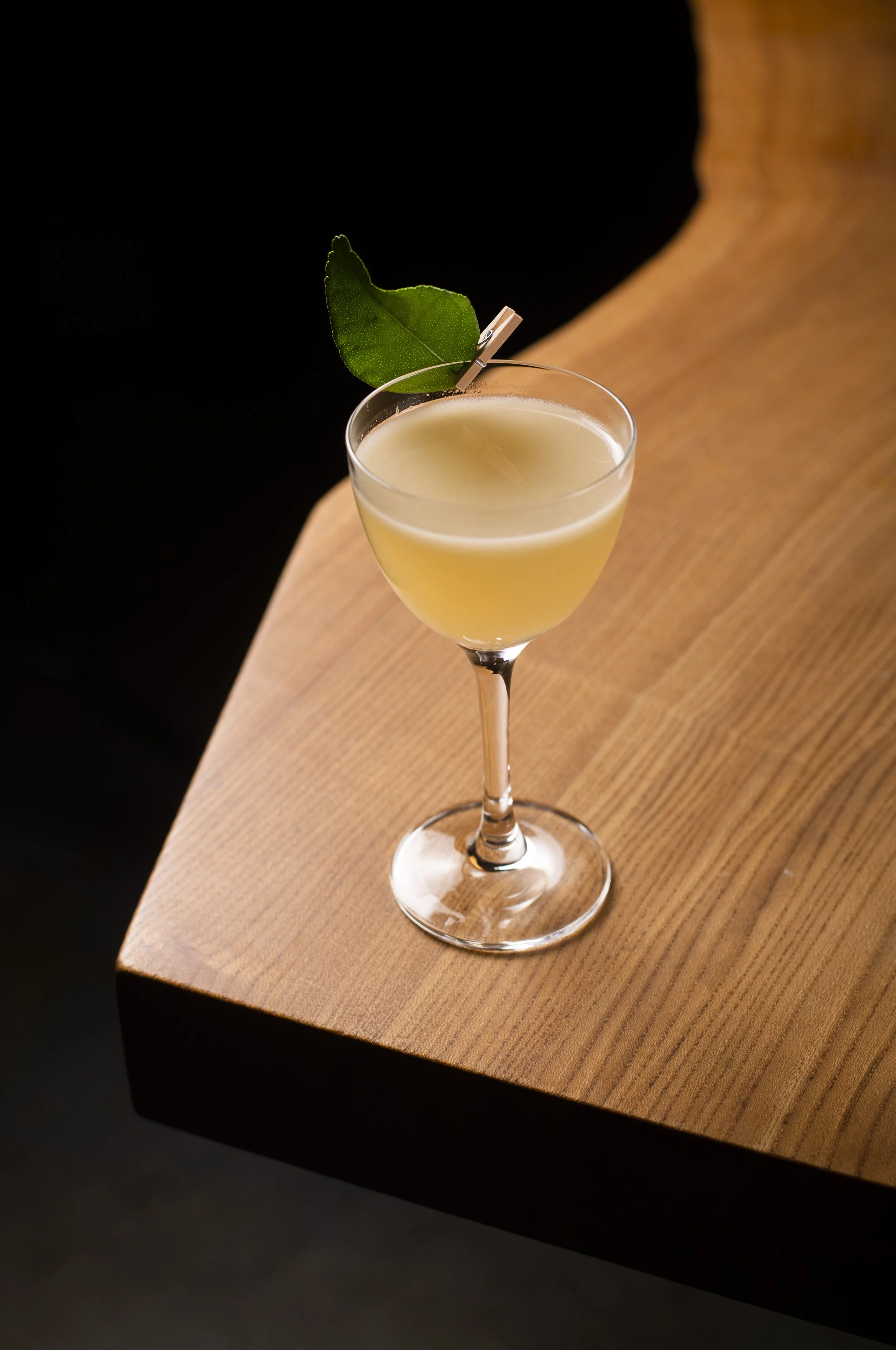 A cocktail in a coupe glass garnished with a green leaf and a small wooden clothespin on a wooden surface.