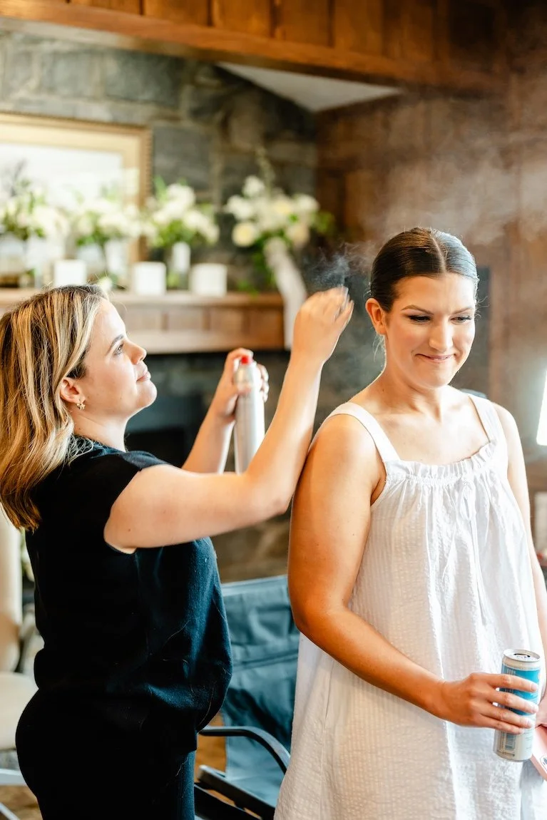 hair stylist dressed in all black spraying hairspray on a bridal hairstyle
