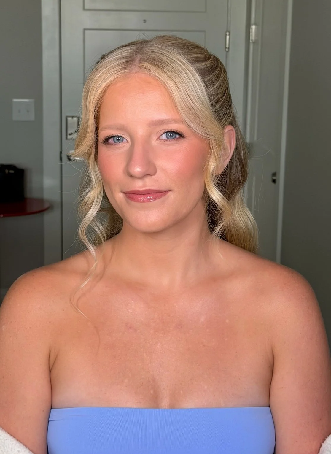 A woman with blonde, wavy hair styled with a half-updo, wearing a strapless blue top, and smiling softly in an indoor setting.