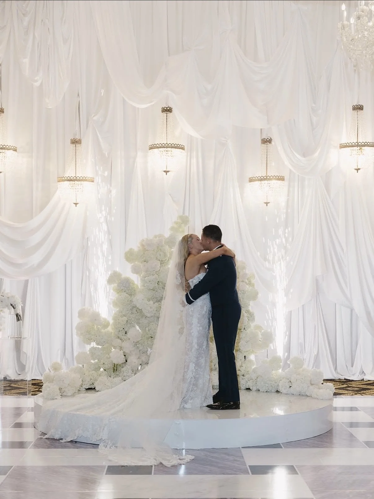 bride and groom kissing in front of all white backdrop and white flowers