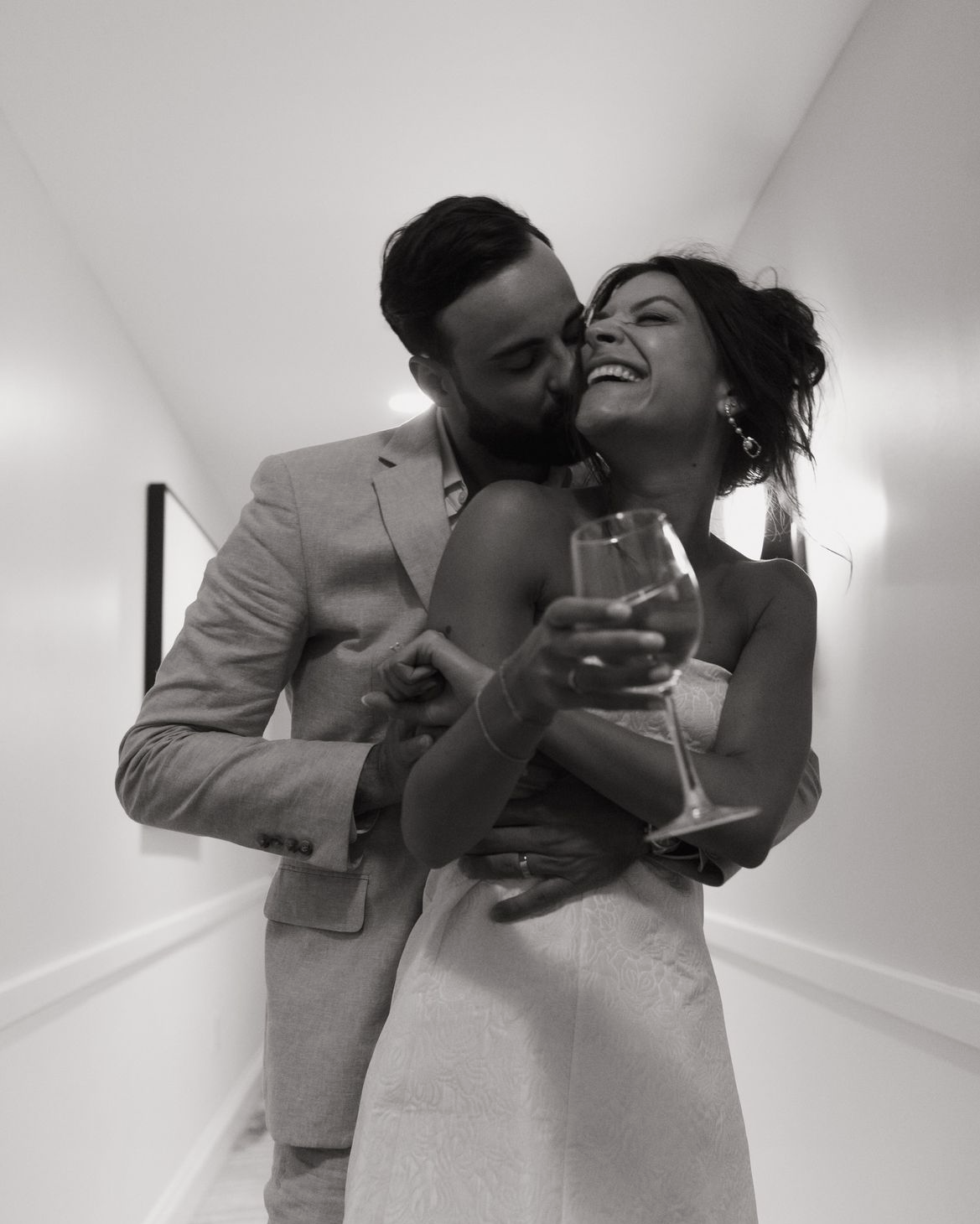 A black and white photo of a joyful couple at a celebration, with the man kissing the woman on the cheek while she holds a wine glass, all in an indoor setting.