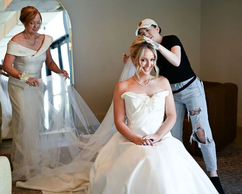 Bride smiling in a white wedding dress, sitting, with two women attending to her in a room.