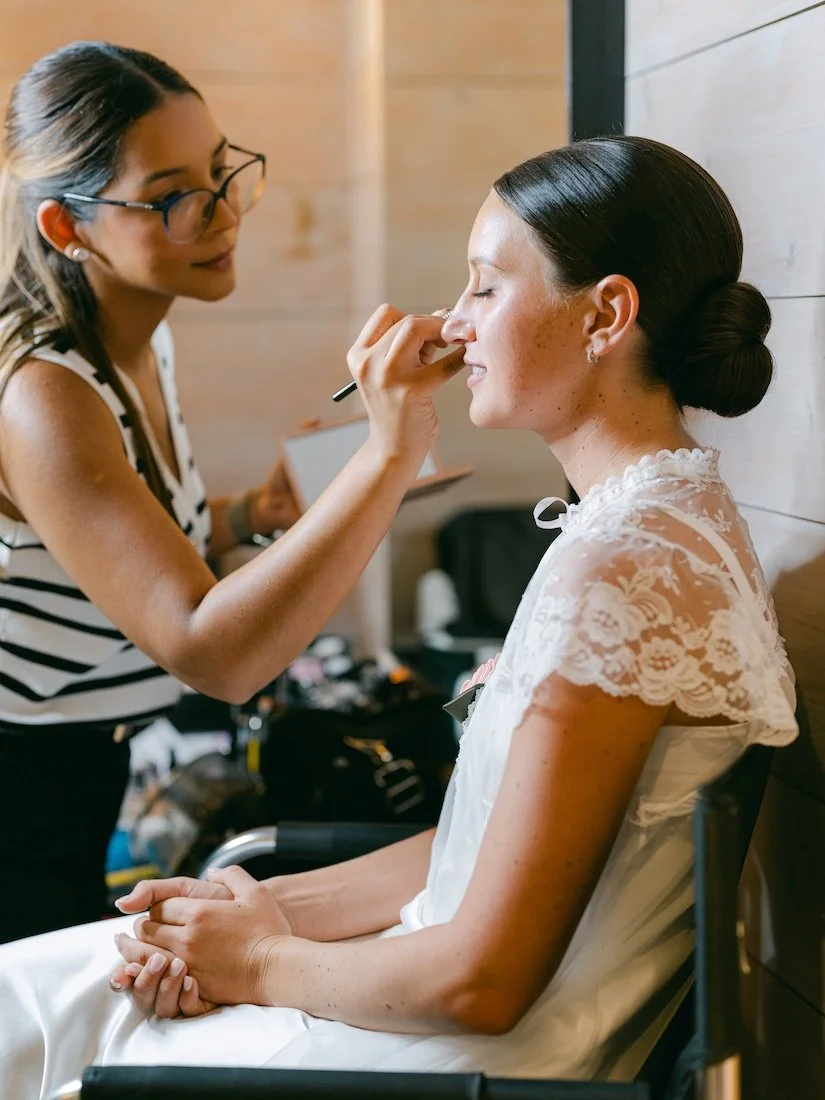 makeup artist wearing glasses and holding a makeup brush as she applies makeup on a brunette bride