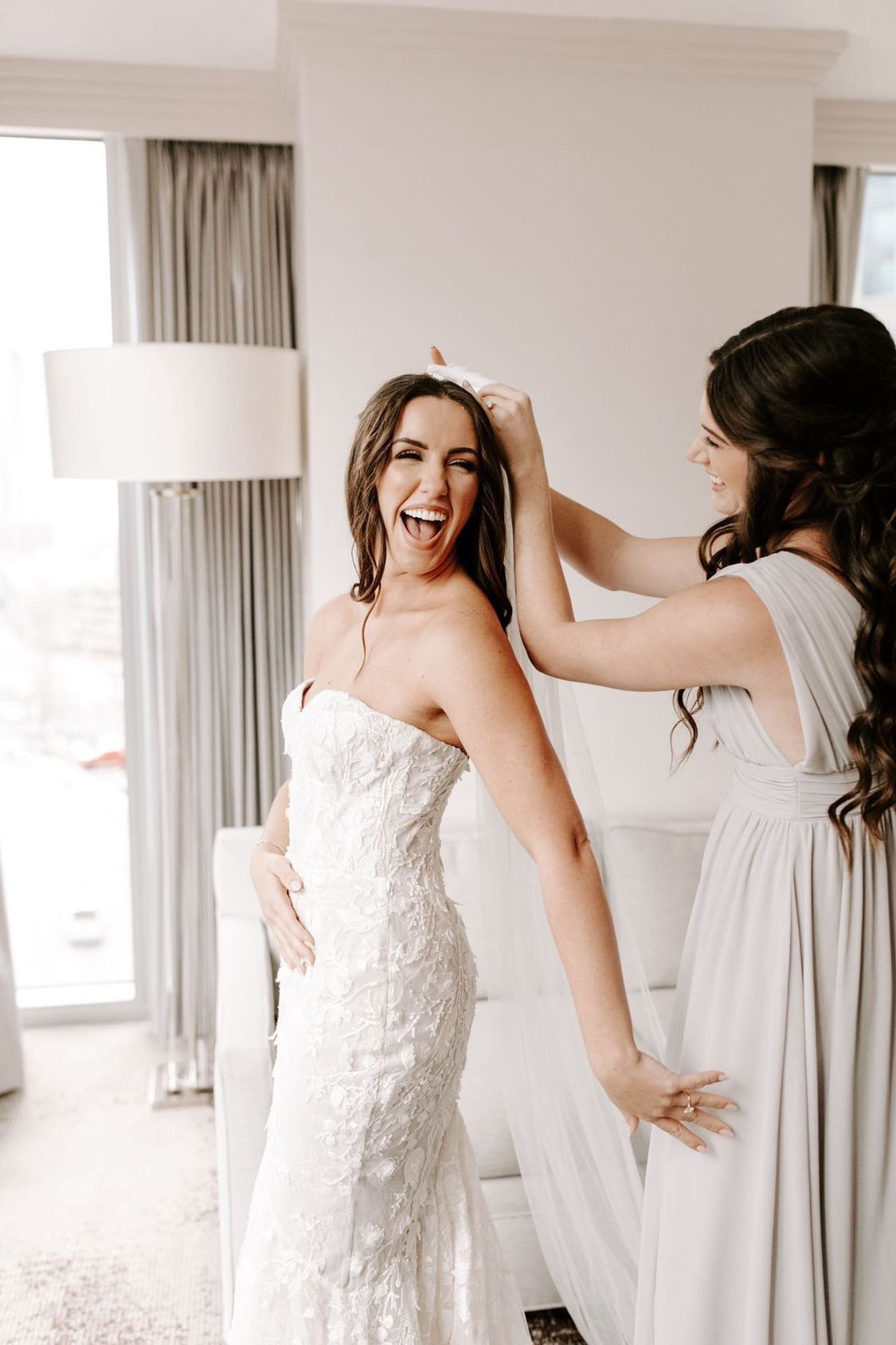 Bride in a strapless lace wedding gown smiling and posing as her friend helps with her dress during a wedding preparation.