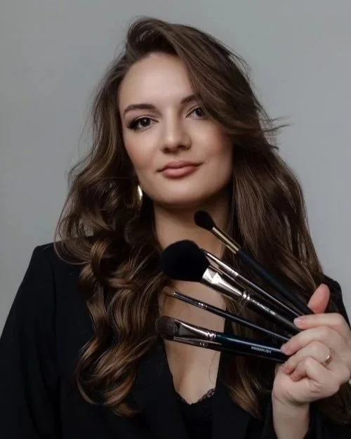 Woman with wavy brown hair holding makeup brushes, dressed in black, against a neutral background.