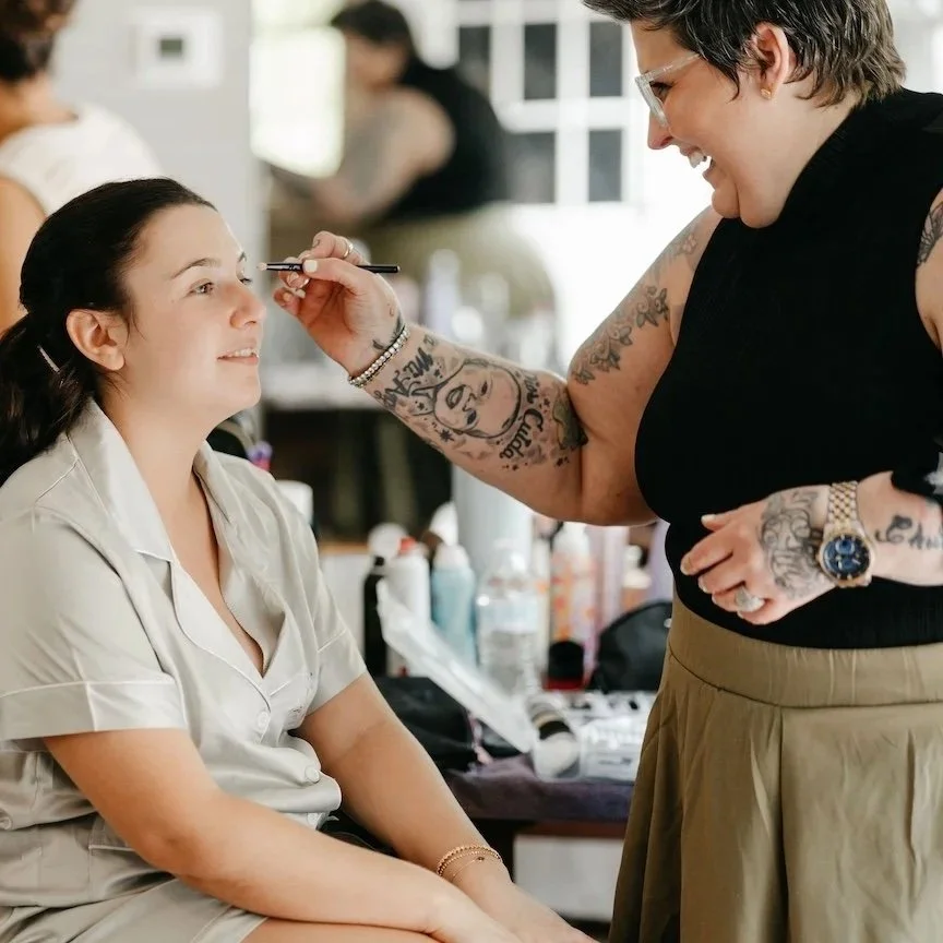 Makeup artist applying eyeshadow to a seated woman with a smile, in a room with makeup tools and products on a table.