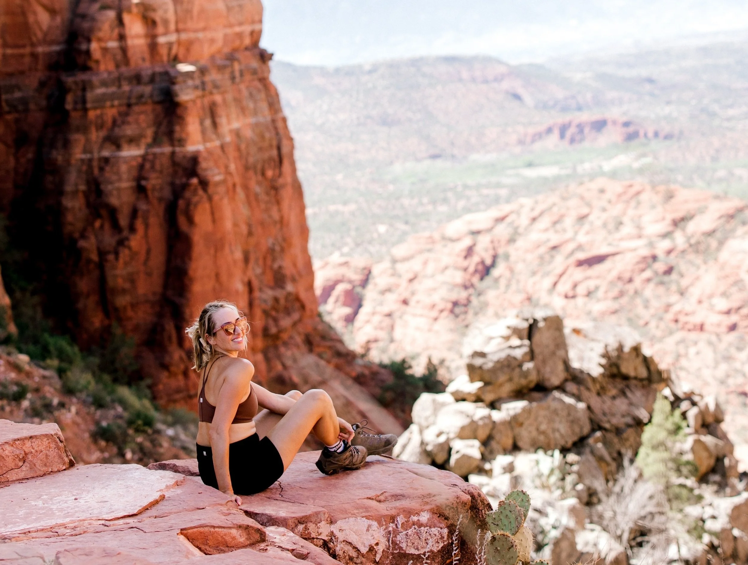 blonde woman sitting on the edge of the mountain turning around to smile with the mountain view in the background