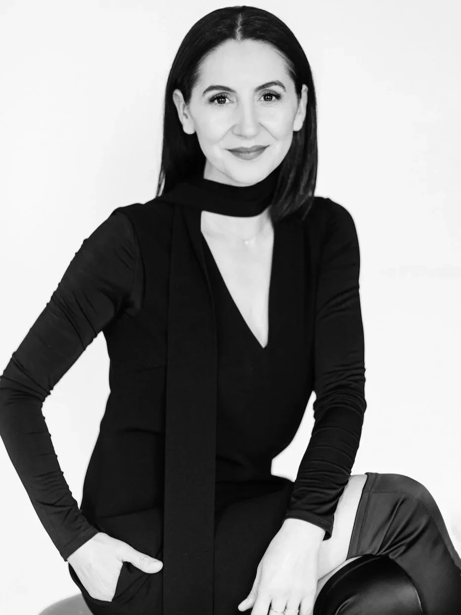 Black and white photo of a woman with dark hair, smiling, wearing a black long-sleeved dress with a matching scarf around her neck, sitting on a stool with one knee bent, with a plain white background.