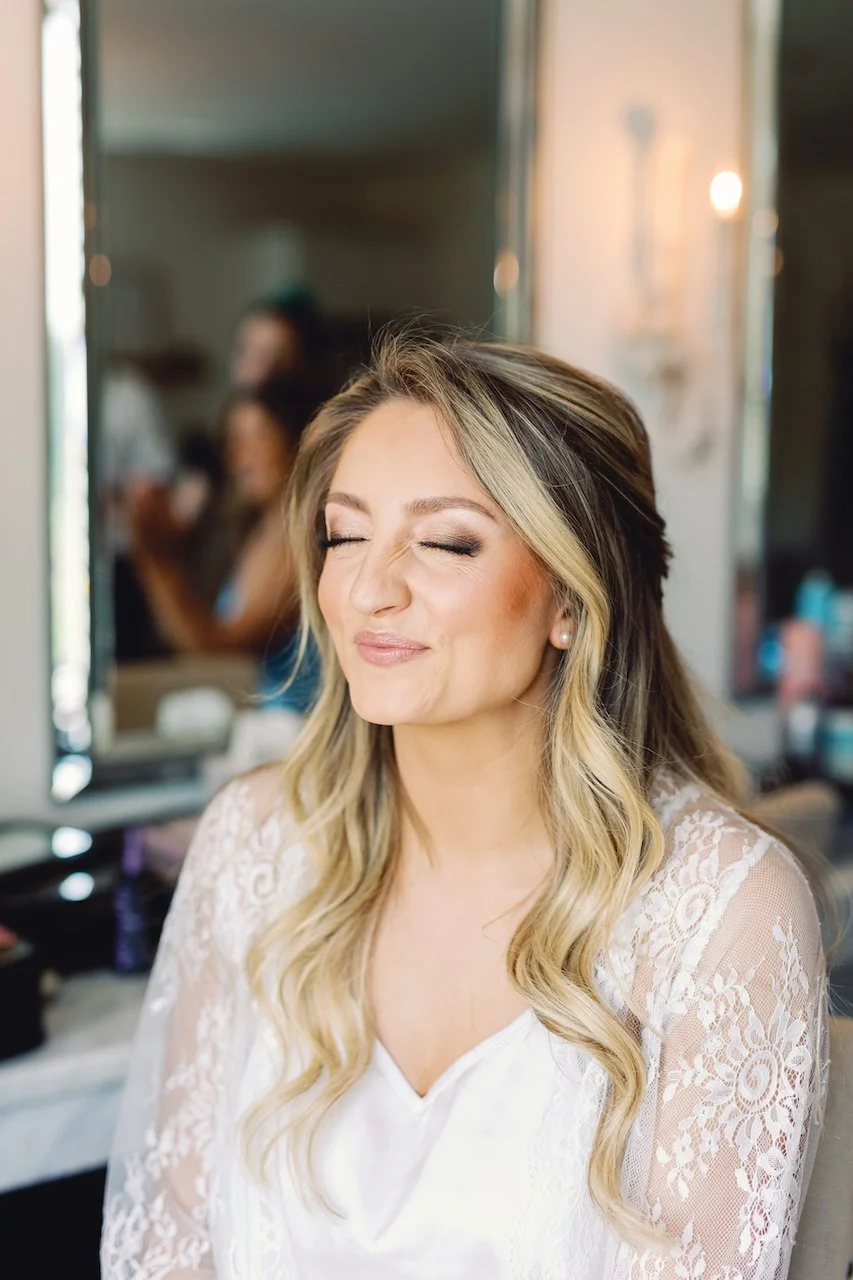Close-up of a smiling woman with long, wavy blonde hair and closed eyes, wearing a white lace top, in front of a mirror in a makeup or dressing room.