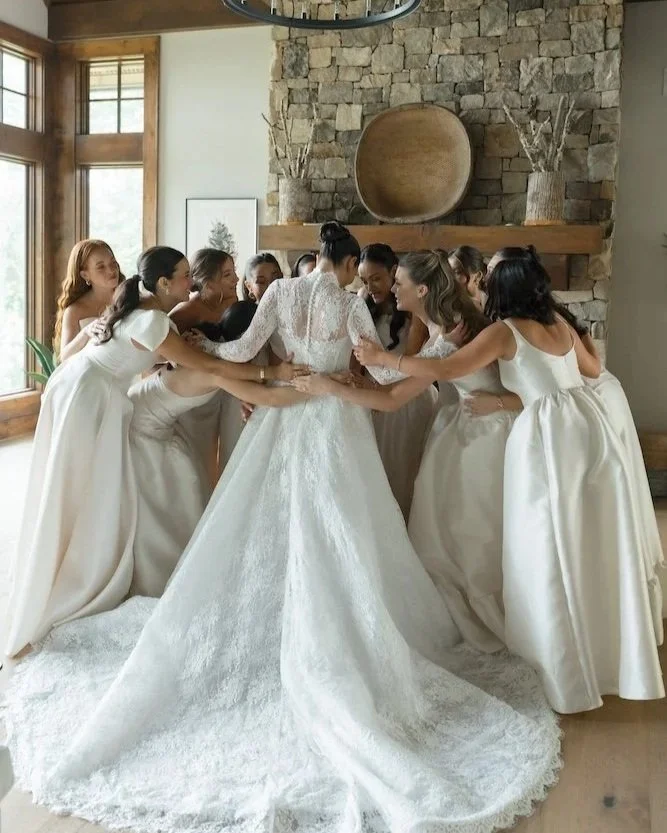 A bride in a white wedding gown surrounded by six bridesmaids in matching white dresses, all holding hands and smiling together inside a room with large windows and a stone fireplace.