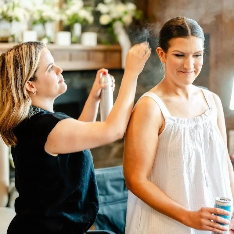 Woman with dark hair in a white sleeveless top smiling as a hairstylist sprays her hair, holding a can of hair spray in her hand, in a cozy indoor setting.