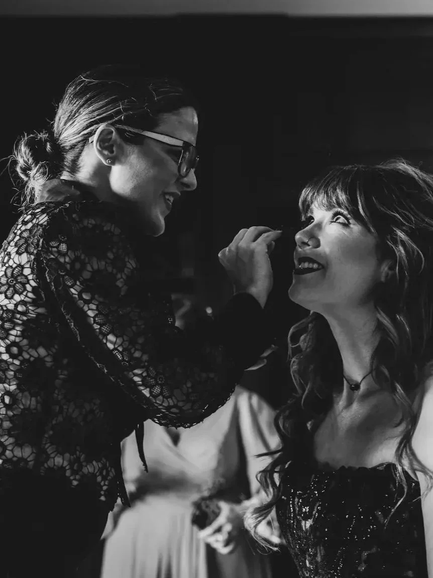 Two women laughing and looking at each other, one applying makeup to the other's face. Black and white image.