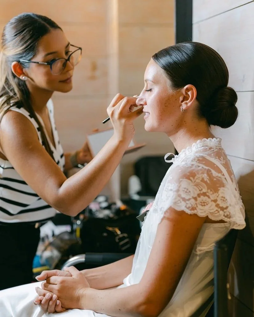 Makeup artist applying makeup to a seated woman with closed eyes in a white lace dress.