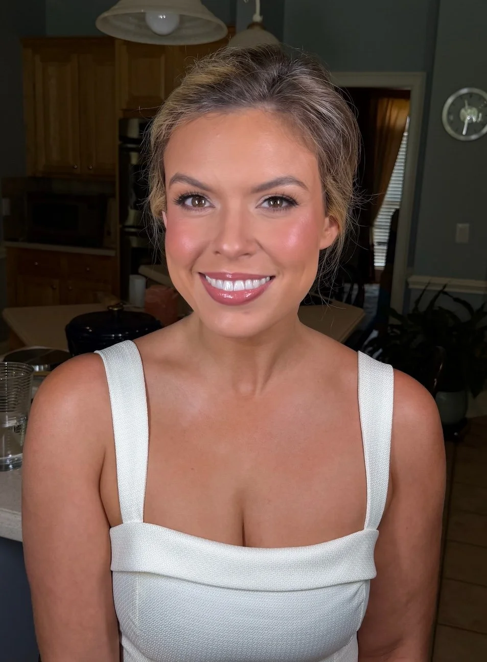 A smiling woman with blonde hair and wearing a white sleeveless dress standing inside a kitchen.