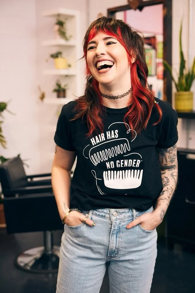 A woman with red and black hair, wearing a black T-shirt with the text "Hair has no gender" and a graphic of two hair combs, standing indoors with a smile.
