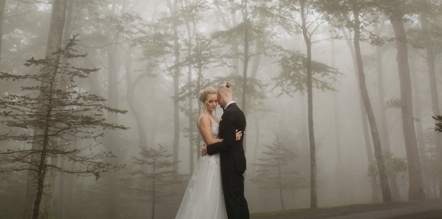 A bride and groom embrace in a foggy forest, the bride wearing a white wedding gown and the groom in a black suit.
