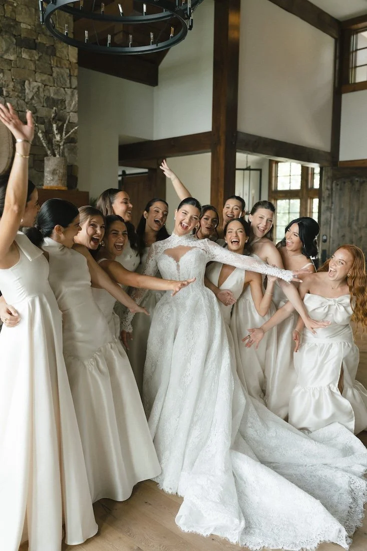 A bride in a white wedding dress surrounded by her bridesmaids in light-colored dresses celebrating happily indoors.