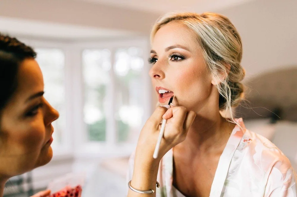 A woman with blonde hair getting her makeup done, while another woman applies lipstick in a bright room with large windows.
