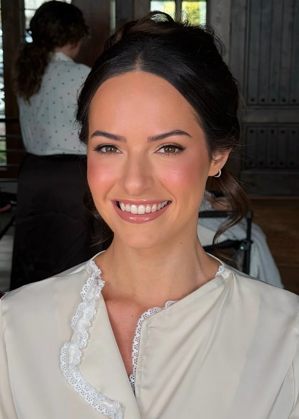 Close-up of a woman with dark hair styled in waves and wearing a cream-colored top with lace detailing, smiling at the camera in a well-lit indoor setting.