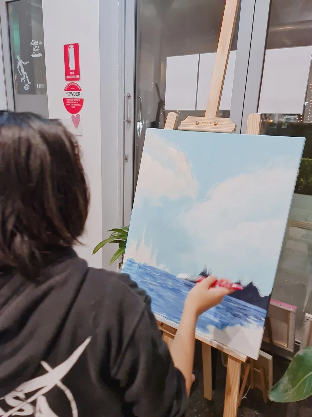 Person painting a seascape on a canvas, depicting sky, clouds, and the ocean, in an indoor art studio.