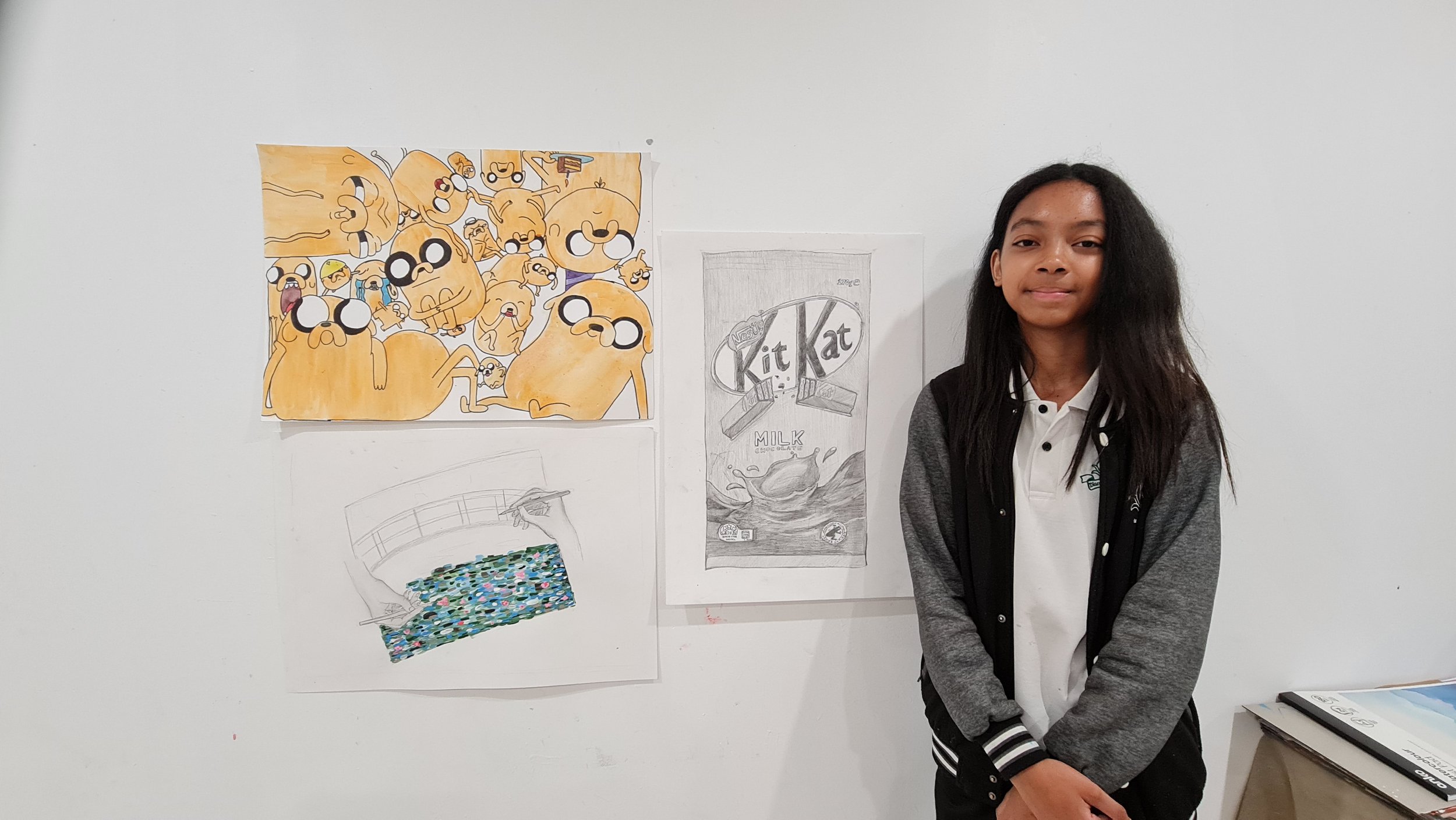 A young girl with long dark hair standing next to a white wall with three posters: a colorful cartoon drawing of various beige animals, a pencil sketch of a Kit Kat chocolate bar with milk splash, and a colored drawing of a pool filled with multicolo
