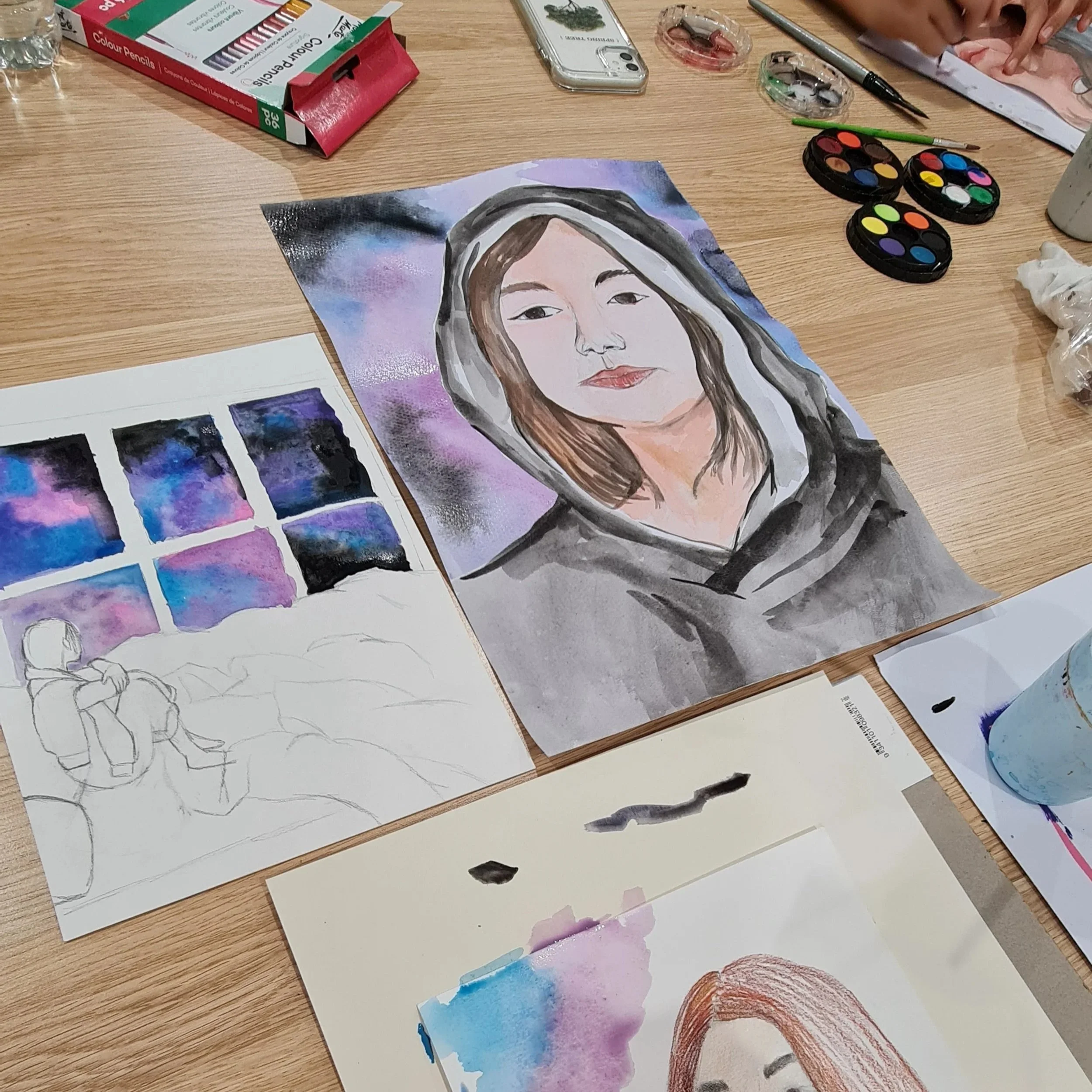 Art supplies on a wooden table, including watercolor paintings, color palette, watercolor paint set, brushes, and a partially completed watercolor portrait of a young woman with long brown hair.