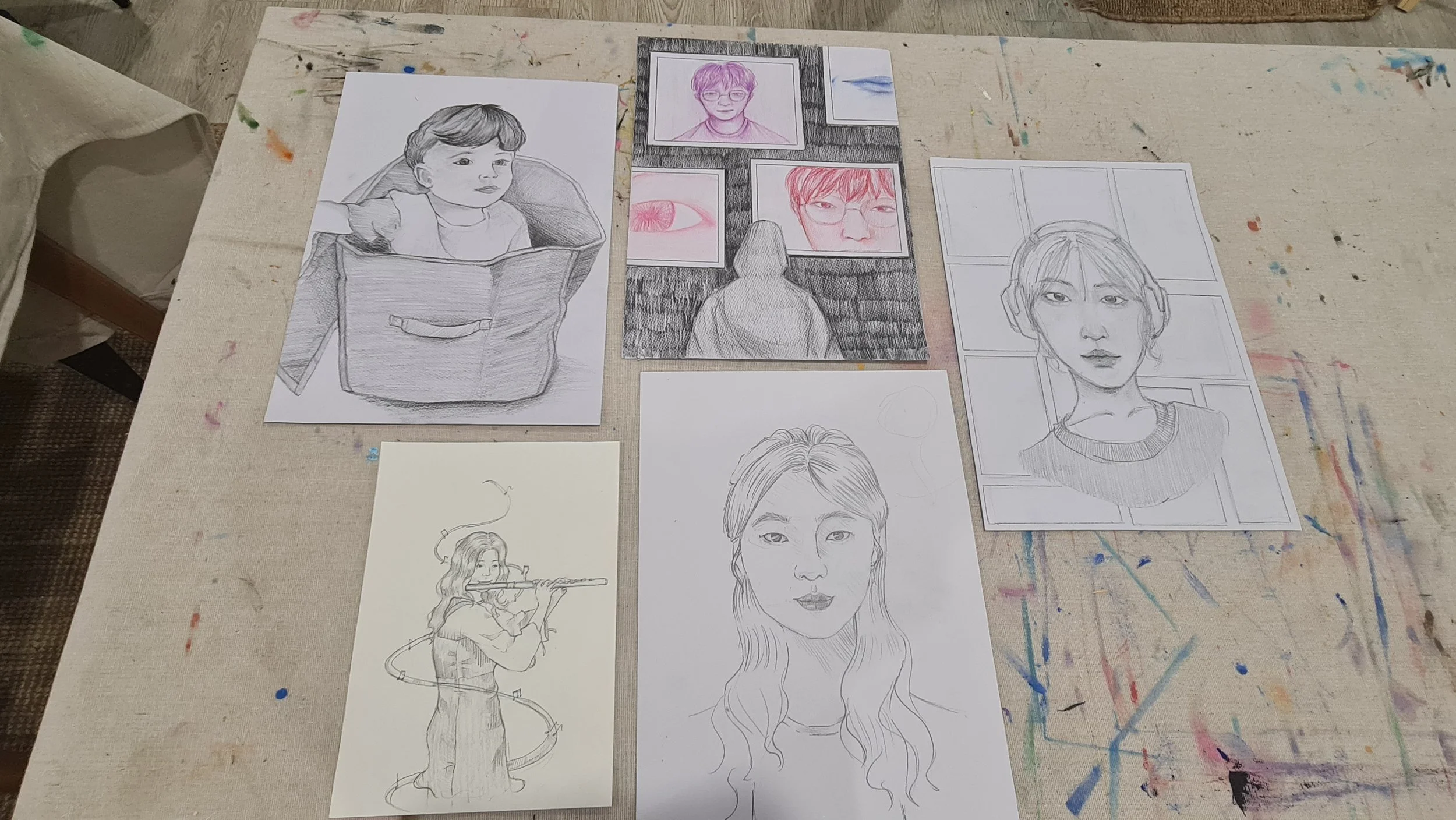 Collection of portrait sketches and drawings of various people, including a child, a woman with headphones, a woman playing the flute, and other individual portraits, spread across a paint-splattered table.