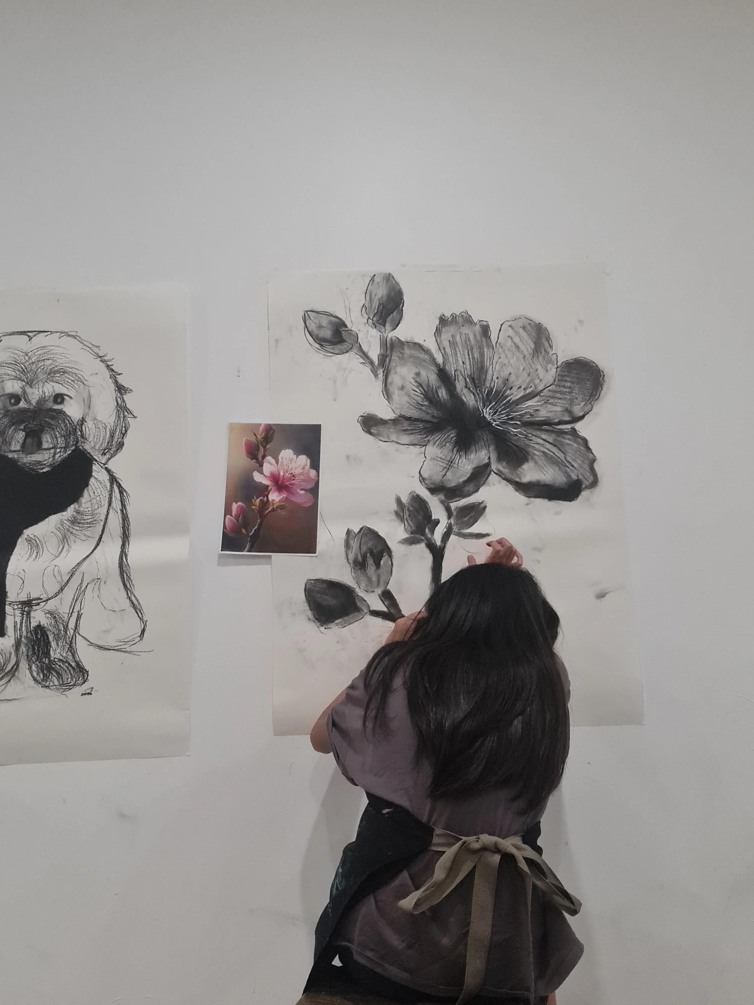 Person with dark hair, wearing a jacket and apron, standing in front of a wall with sketches of a puppy and a floral illustration, engaging in drawing or painting.
