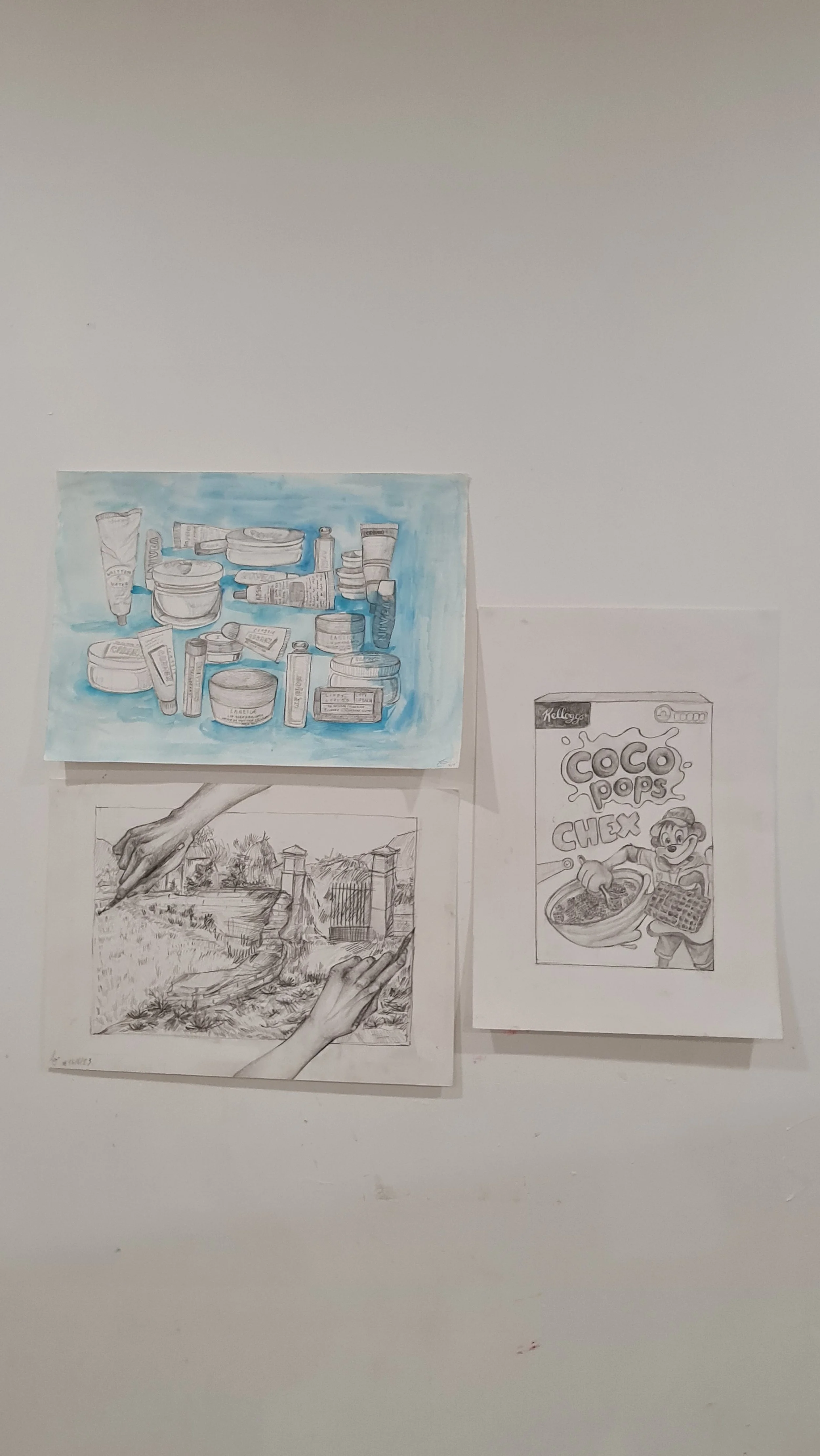 Collection of hand-drawn pictures on paper pinned to a white wall, including bottles and jars, a landscape with trees and a fence, and a comic-style drawing of a cereal box with a cartoon character.