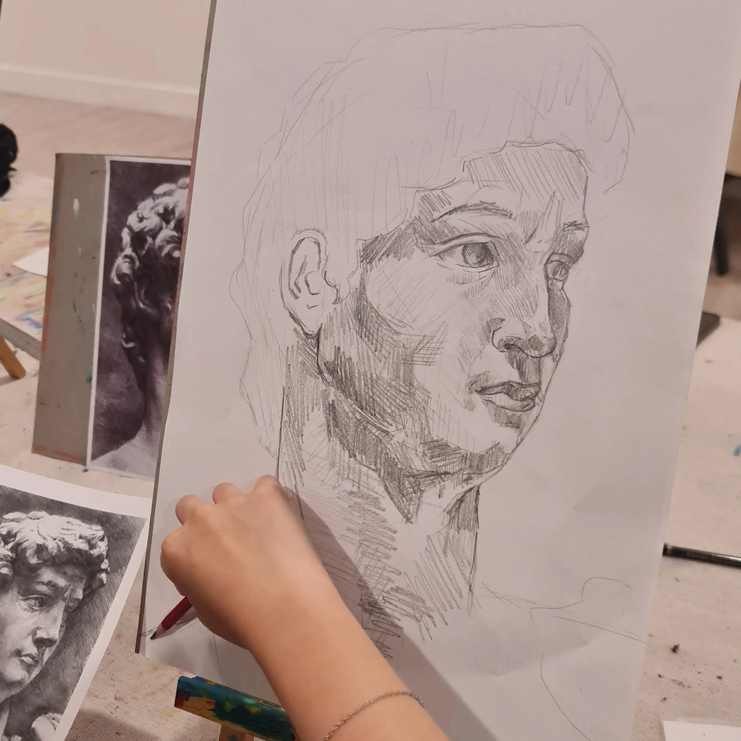 A hand holding a pencil sketches a detailed portrait of a person's face on a piece of paper, with photo references in the background.
