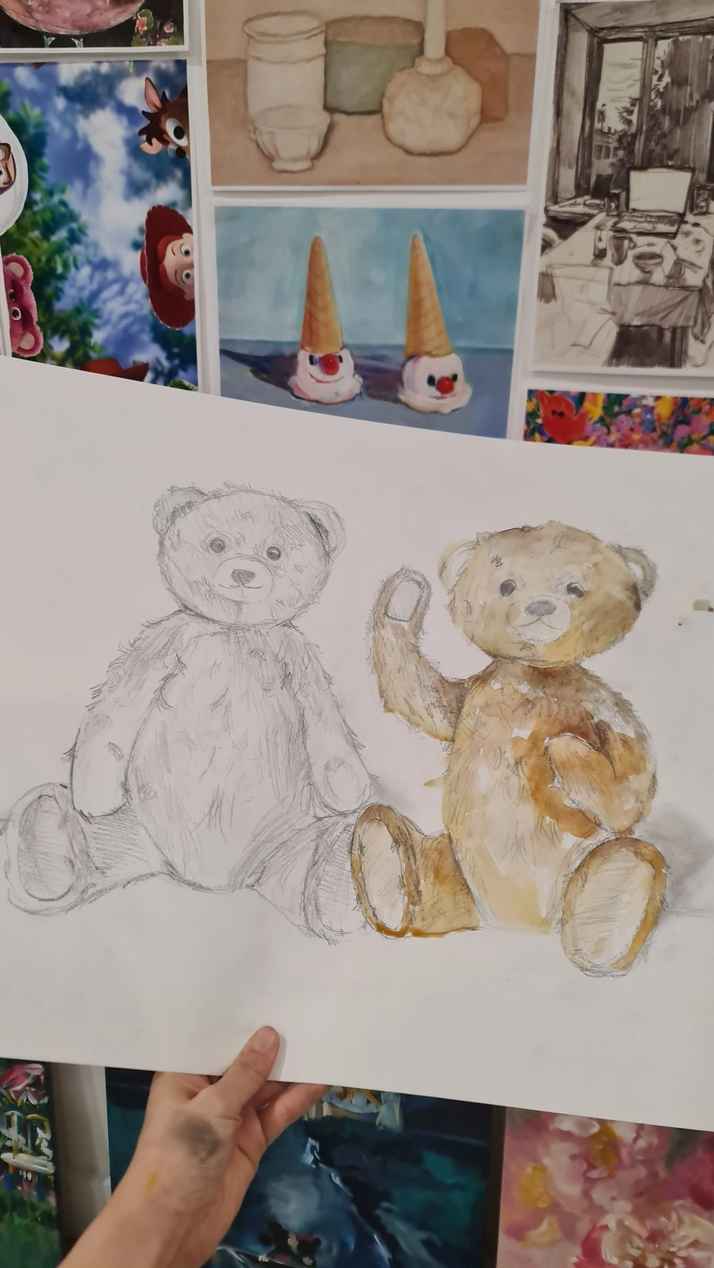 A hand holding a sketch of a teddy bear, with part of the teddy bear painted in color on the right side. In the background, various colorful illustrations and sketches are visible on a wall.