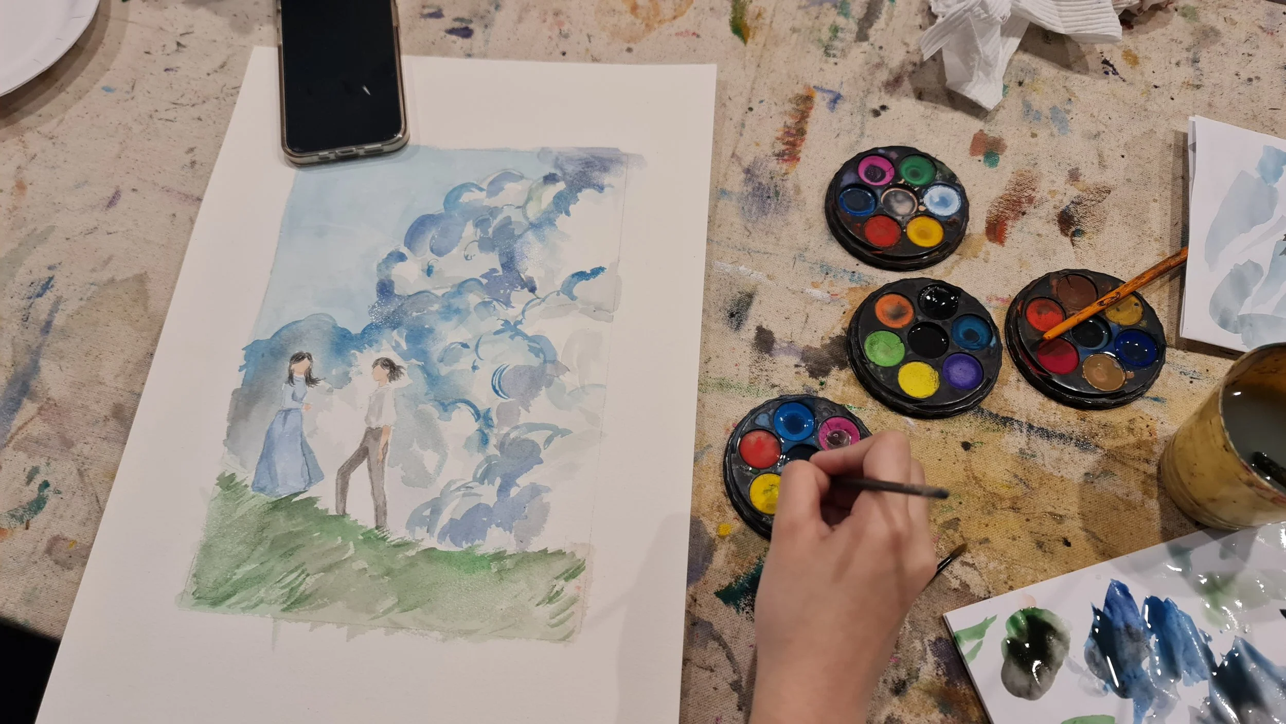 An artist painting a watercolor of two people standing outdoors with clouds and trees, surrounded by watercolor paints, brushes, and art supplies on a paint-splattered table.