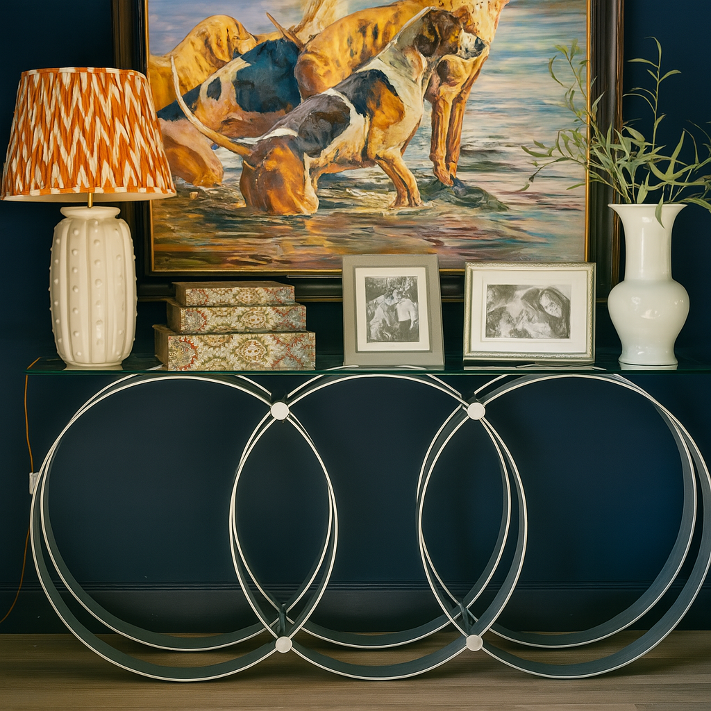 Elegant console table with overlapping circular metal supports and glass top, styled against a navy blue wall with framed dog painting, patterned lamp, floral boxes, black-and-white photos, and white vases with foliage on light wood flooring.
