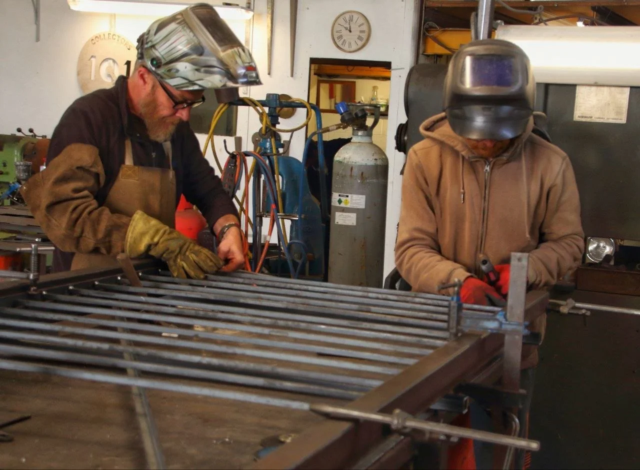 Two welders assembling a bespoke mild steel gate in a well-equipped workshop. Both wear protective helmets and gloves while working on a clamped metal frame atop a heavy-duty workbench.