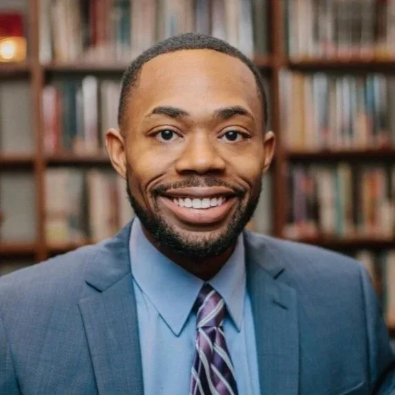 A male attorney dressed in a blue suit with a striped tie, smiling, standing in front of bookshelves.