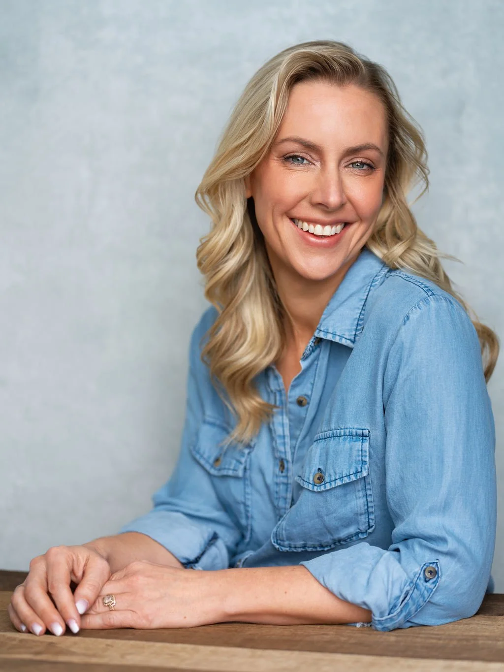 Ally Murphy is smiling at camera with her hands on a table, wearing a denim blue shirt.