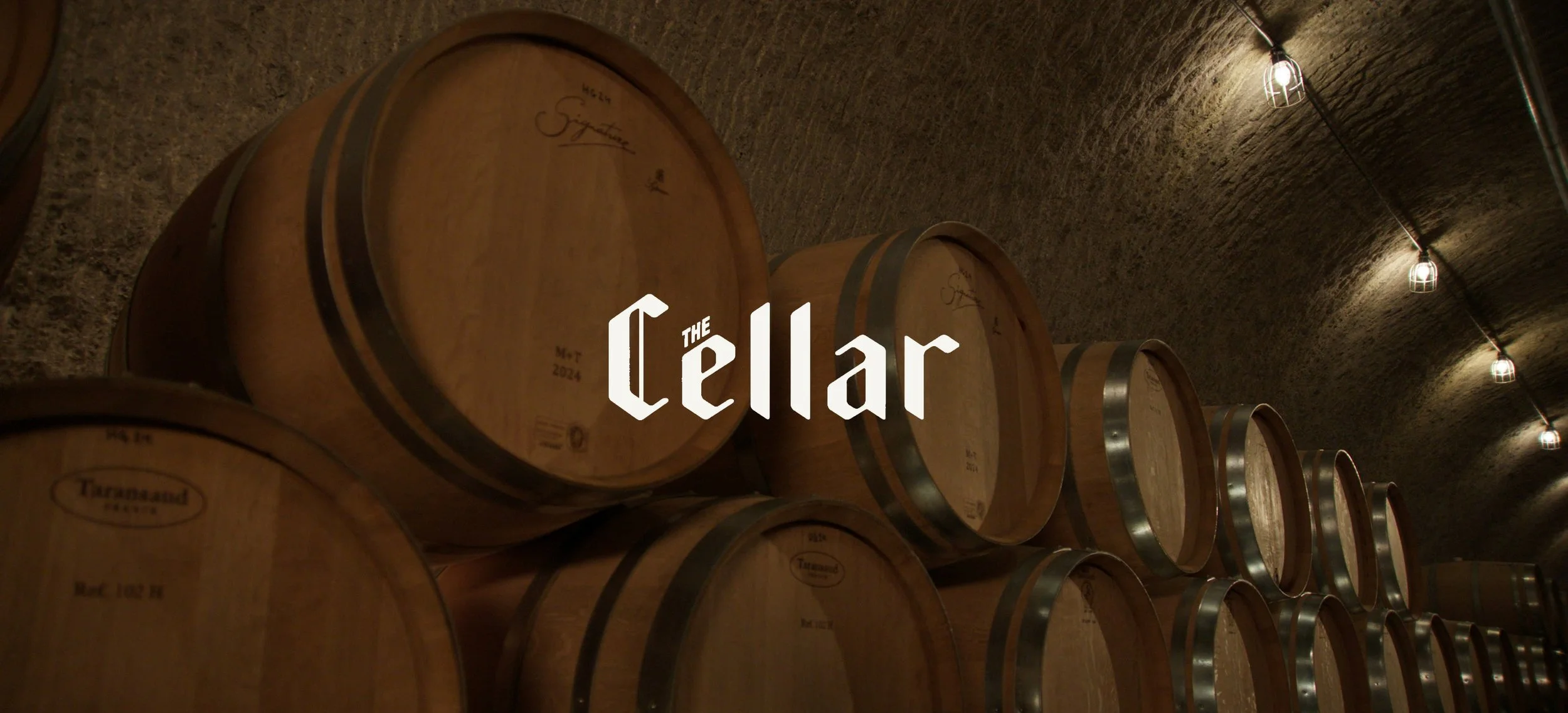 Wine barrels stacked in a cellar with the words "The Cellar" centered in white letters.