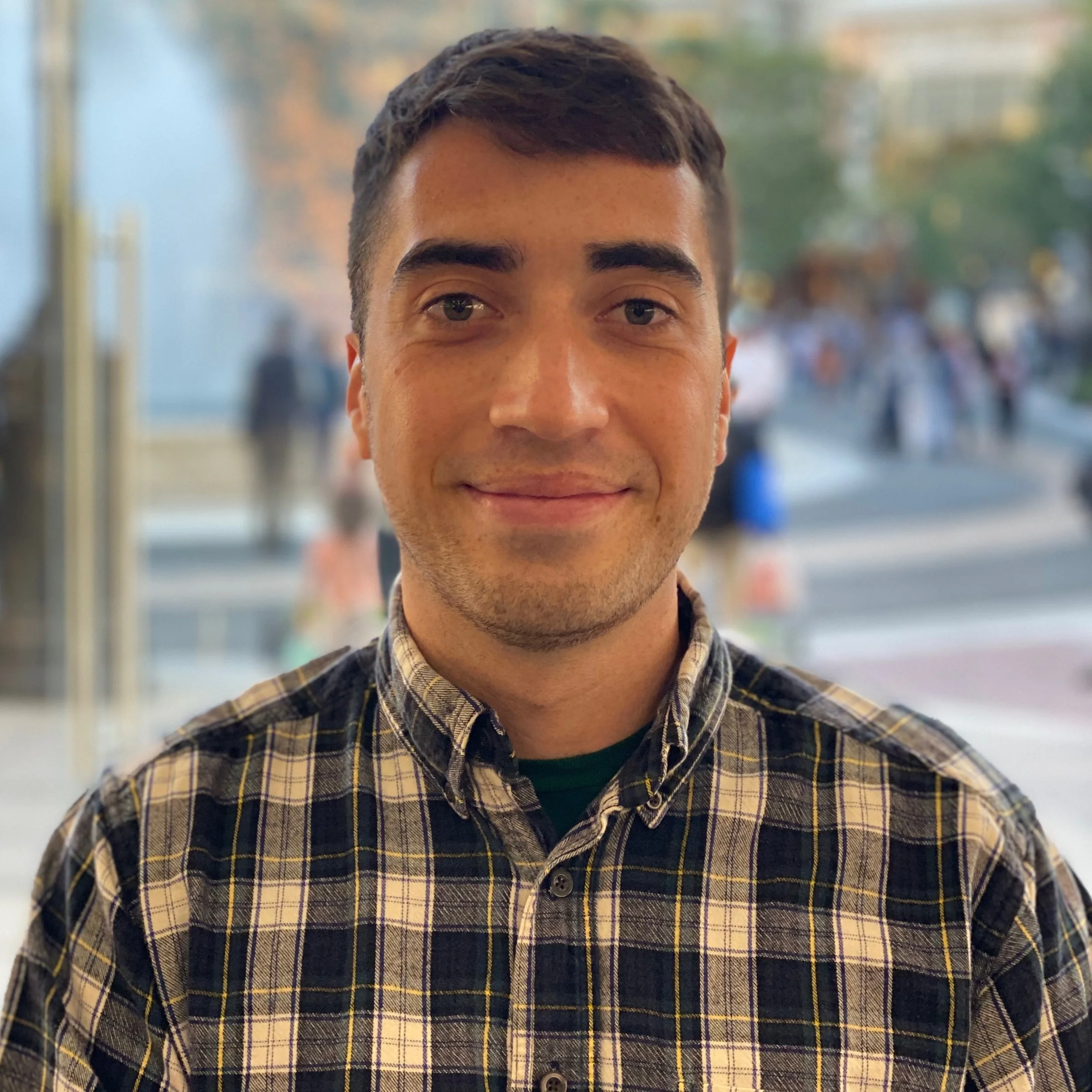 Close-up of a young man with short dark hair, fair complexion, wearing a plaid shirt, smiling indoors with a blurred outdoor scene in the background.