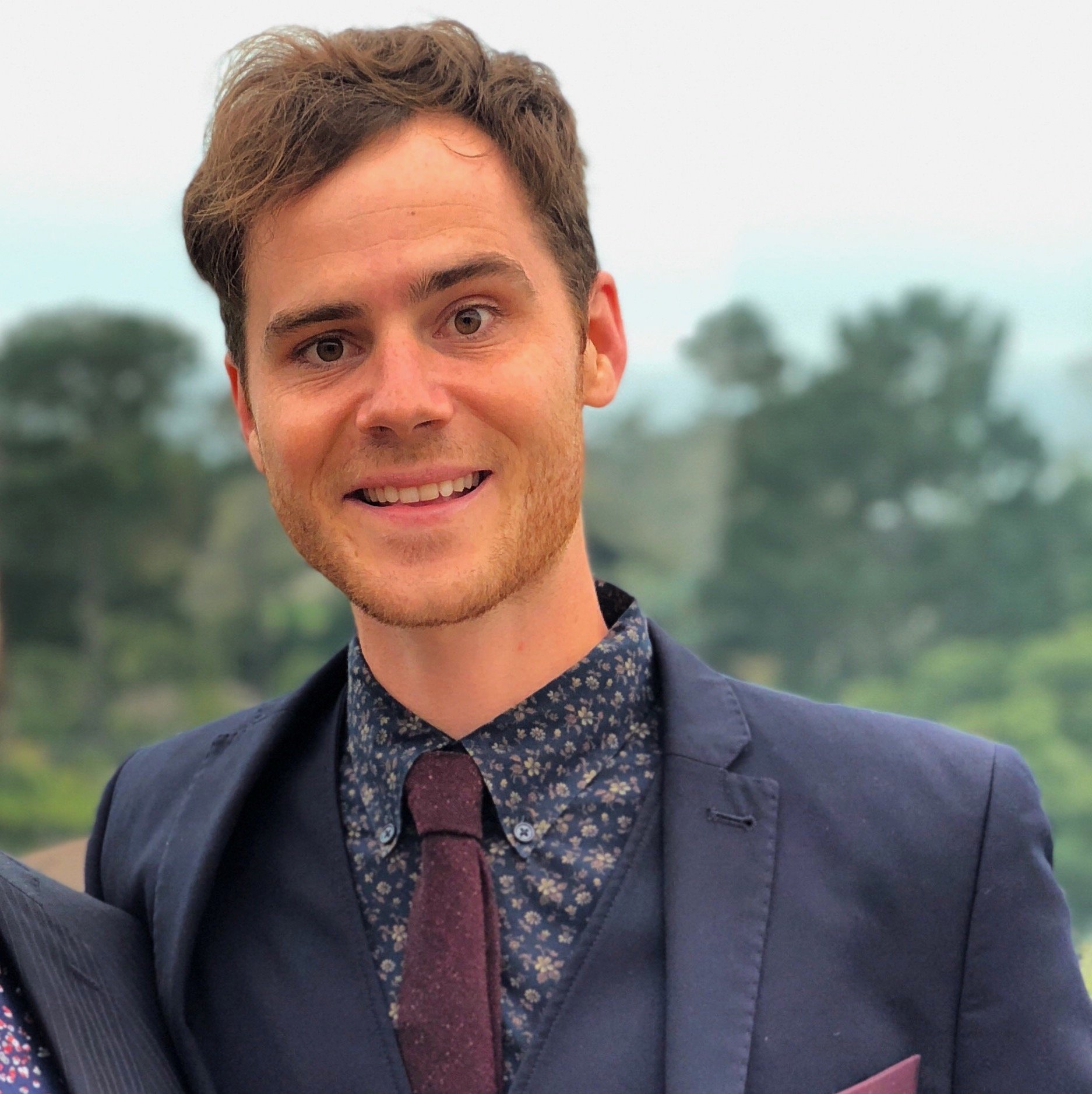 Close-up of a smiling young man with brown hair, wearing a navy blazer, patterned shirt, and maroon tie, outdoors with blurred trees in the background.