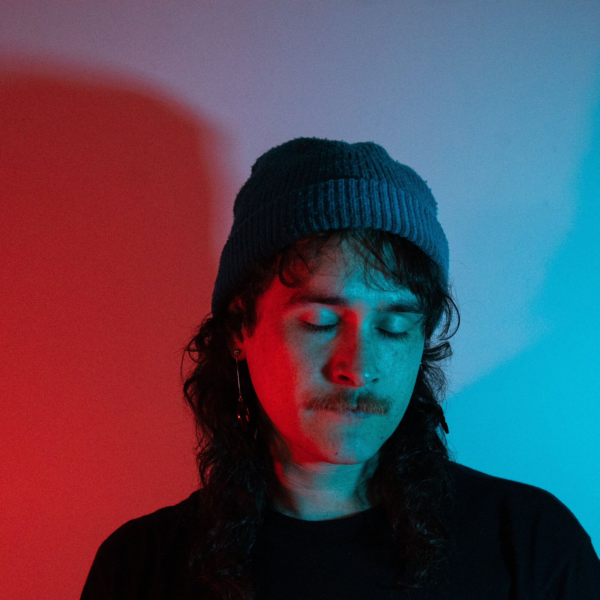 Teenager with long hair, mustache, closed eyes, wearing a gray beanie, illuminated by red and blue lights.