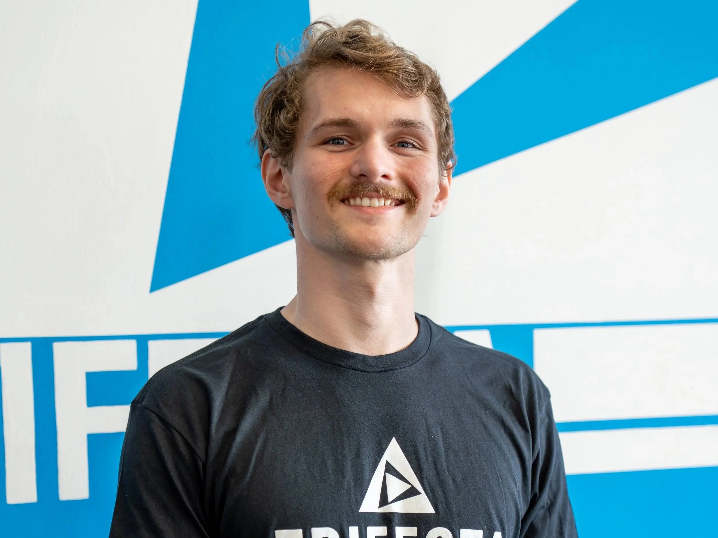 Young man with curly blond hair and a mustache smiling, wearing a black T-shirt with a triangle logo, standing in front of a blue and white background with the words 'TRIFECTA' visible.
