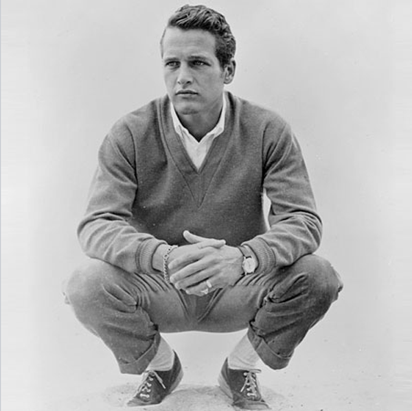 Black and white photograph of a young man squatting with hands clasped, wearing a sweater, pants, sneakers, and a watch.
