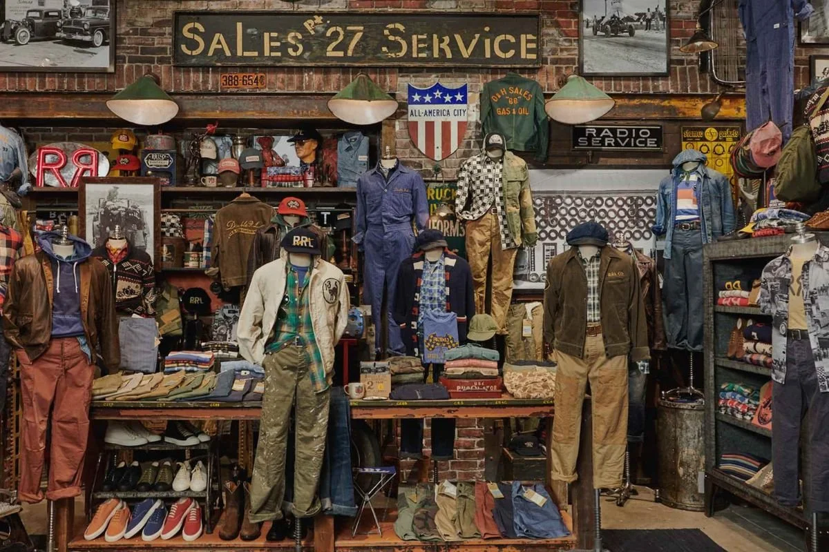 Clothing store with mannequins dressed in vintage apparel, hats, and accessories, brick wall background with signs and framed photos, indicating a retro or thrift shop.